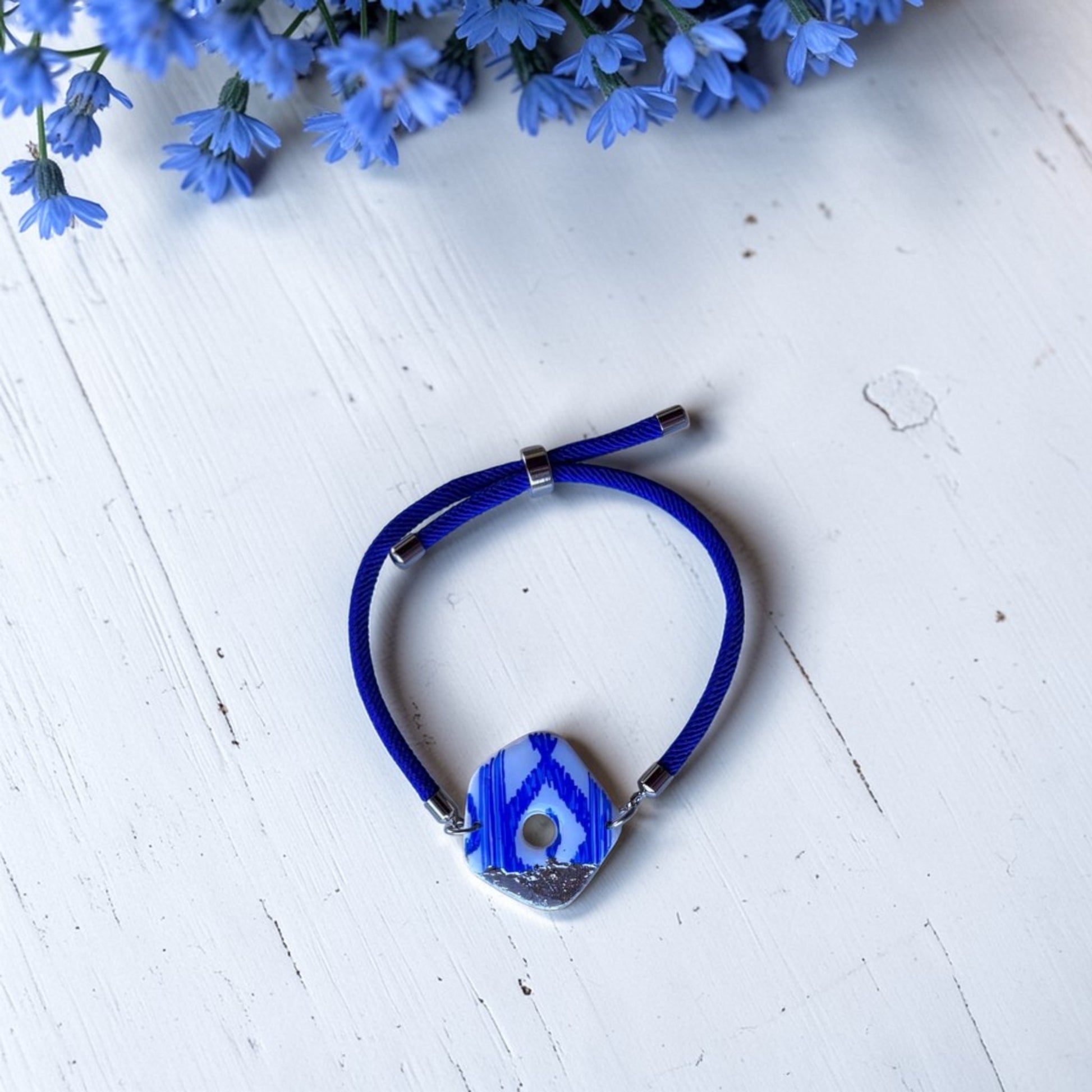 Pulsera artesanal con pieza central de arcilla polimerica con estampado de lenguas mallorquinas pintado a mano, azul y blanca sobre cordón náutico azul. Fondo de madera blanca y flores azules en la parte superior de la imagen.