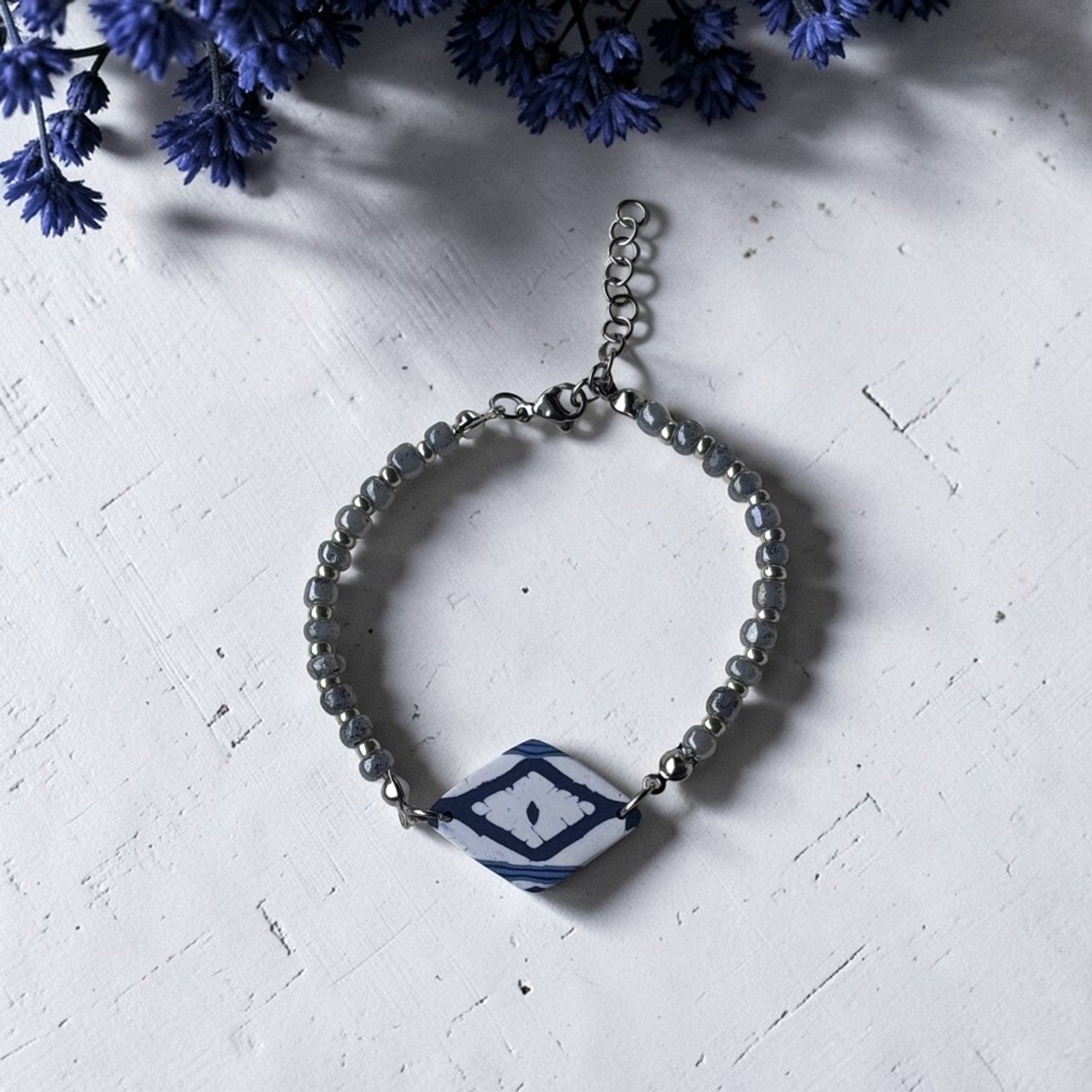 Pulsera artesanal Llengos Sal con pieza central rombo de arcilla polimérica en azul marino y blanco y cuentas de cristal azul claro plateado con cierre regulablede acero inoxidable, sobre fondo blanco con flores azules. Colección Esencia Mallorquina de Pm_Art.
