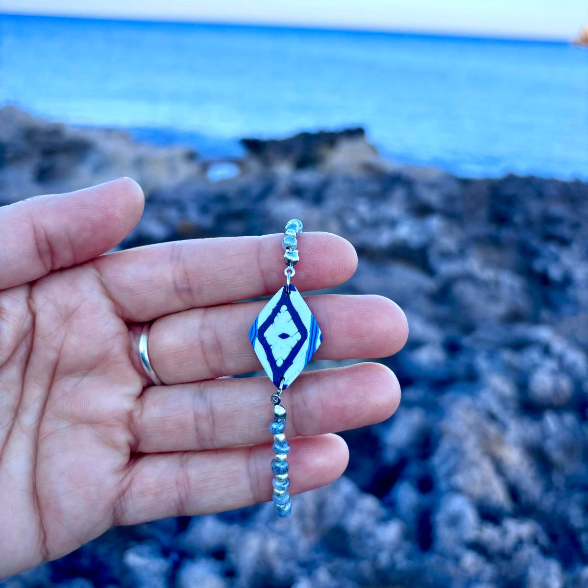 Pulsera artesanal Llengos Sal con pieza central rombo de arcilla polimérica en azul marino y blanco y cuentas de cristal azul claro y plateado, sostenida en mano con la costa rocosa y el mar Mediterráneo de Mallorca al fondo. Colección Esencia Mallorquina de Pm_Art.
