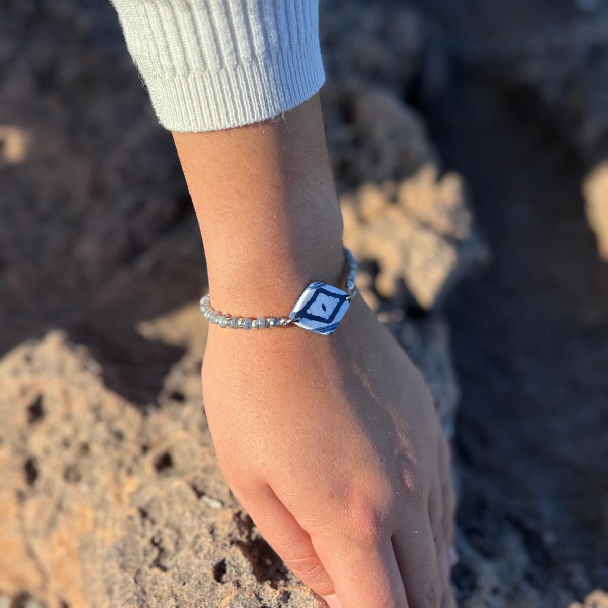 Pulsera artesanal Llengos Sal con pieza central rombo de arcilla polimérica en azul celeste y marino y cuentas plateadas, puesta en muñeca sobre roca natural de Mallorca. Colección Esencia Mallorquina de Pm_Art.