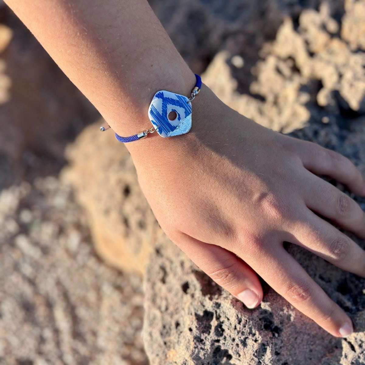 Pulsera artesanal de la colección Esencia Mallorquina lucida en la muñeca. Destaca una pieza central de arcilla polimérica con estampado de lenguas azules y blancas sobre un cordón náutico azul ajustable, con fondo de rocas marinas.