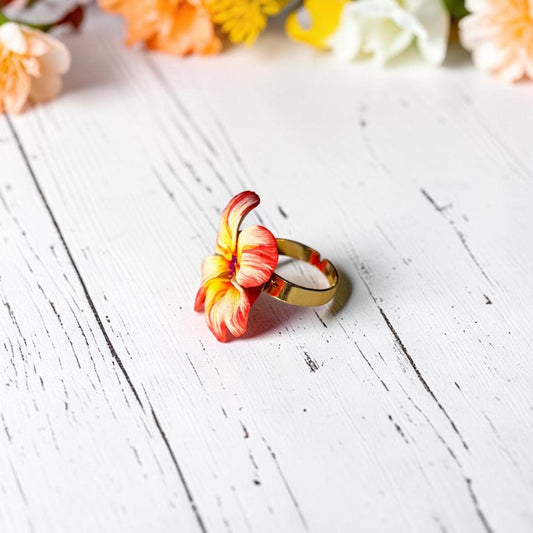 Vista lateral anillo flor, de arcilla polimérica, hecho a mano, en tonos naranja combinado con blanco y amarillo, centro de color morado, sobre madera blanca envejecida