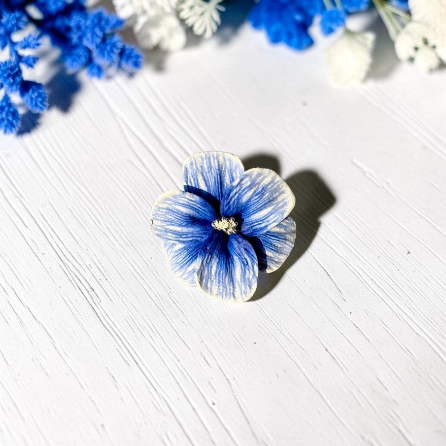 Anillo flor de arcilla polimérica en color azul y blanco. Vista frontal sobre madera blanca y flores azules
