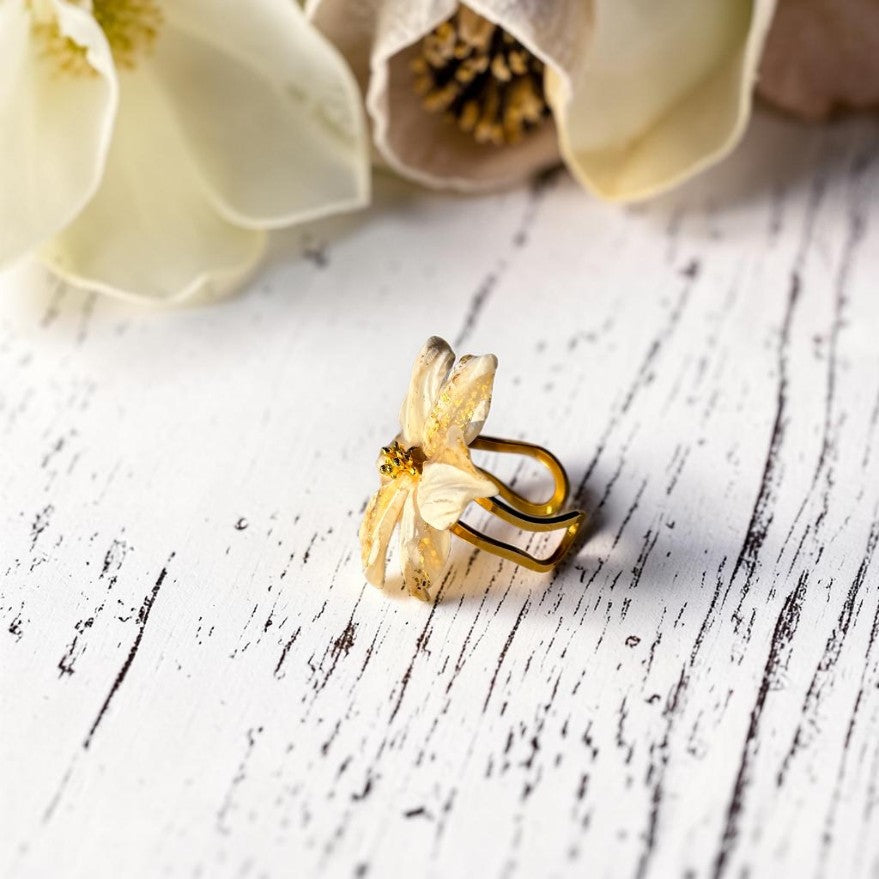 Vista lateral de anillo flor en tonos dorado, beig y blanco hecho a mano, base de acero inoxidable color dorado.