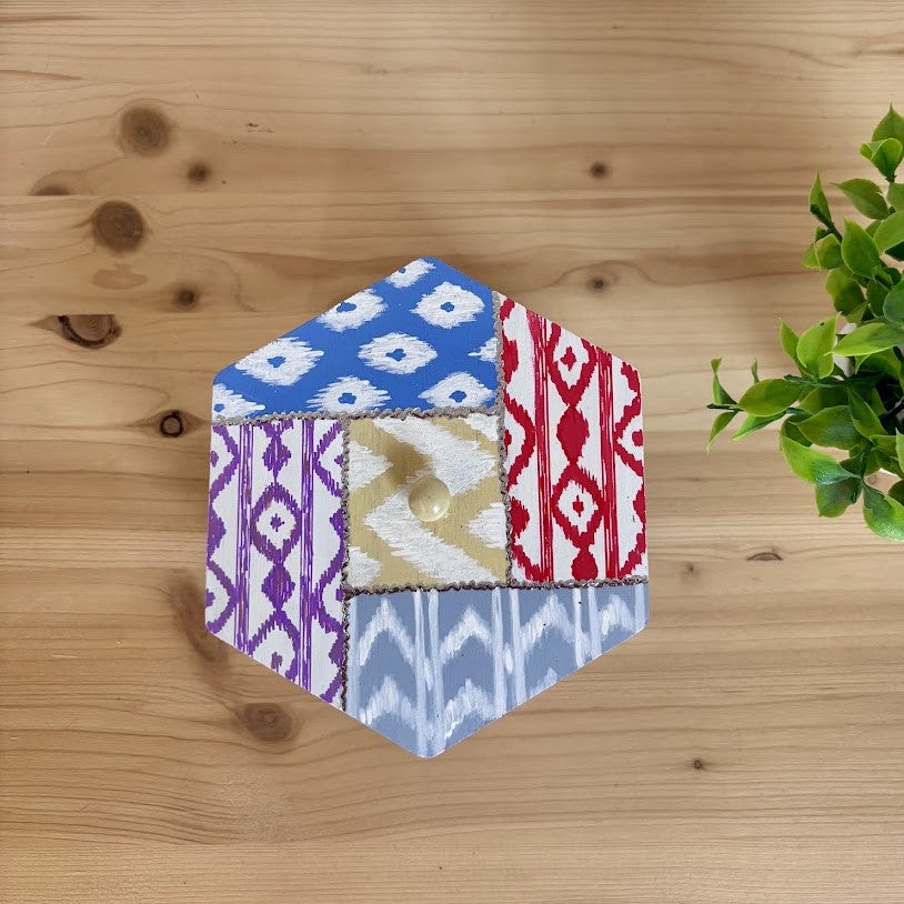 Vista superior de una caja decorativa hexagonal con base metálica blanca y tapa de madera pintada con motivos de la artesanía textil de Mallorca.