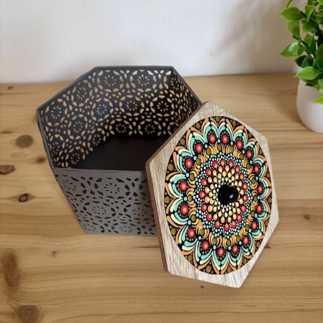 Caja calada de metal gris con su tapa de madera apoyada, decorada con un detallado mandala hecho a mano en puntillismo de colores cálidos y turquesa