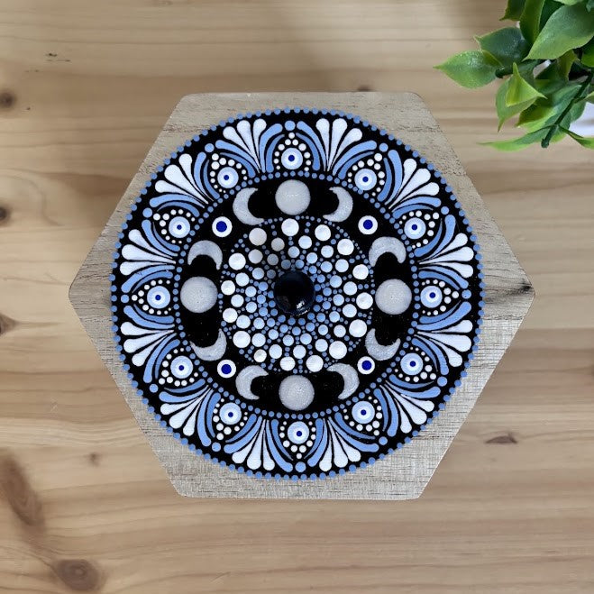 Plano aéreo de la tapa de madera hexagonal pintada a mano con un mandala de fases lunares en tonos azul, blanco y negro con técnica de puntillismo y pomo central.