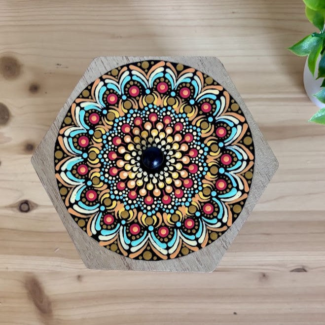 Imagen aérea de la tapa de madera hexagonal para joyero con un intrincado mandala de puntillismo en tonos naranja, turquesa, amarillo y detalles dorados.
