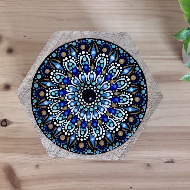 Tapa hexagonal de madera pintada a mano con un diseño de mandala en círculos concéntricos de puntillismo azul, turquesa, blanco y oro.