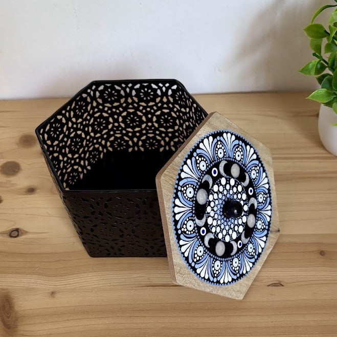 Caja metálica hexagonal negra con calados decorativos y tapa de madera abierta pintada a mano con diseño de fases lunares y puntillismo azul y blanco