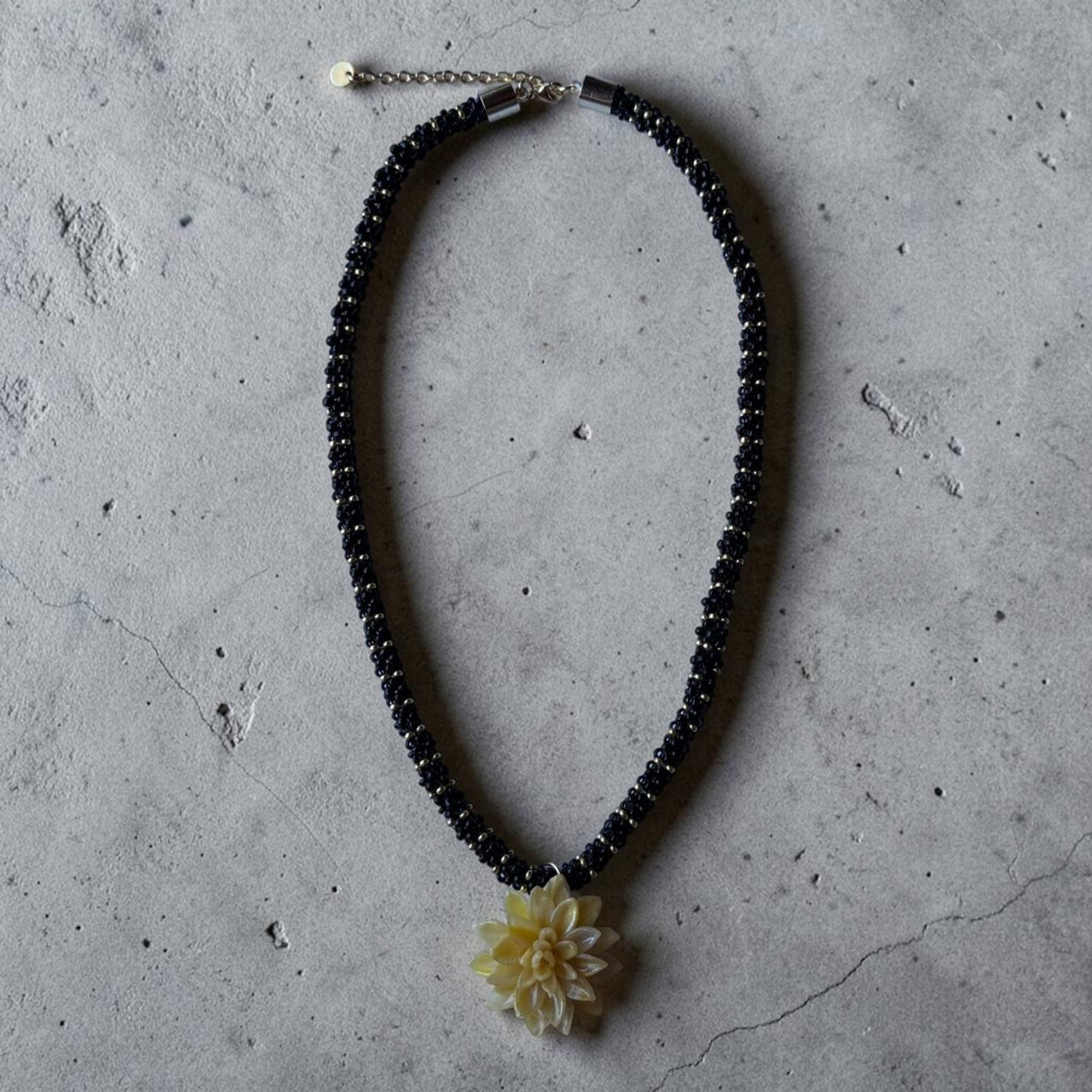 Collar Galatzó con colgante de flor amarilla y cordón  tejido a mano con cuentas de vidrio negras y detalles dorados sobre fondo de cemento.