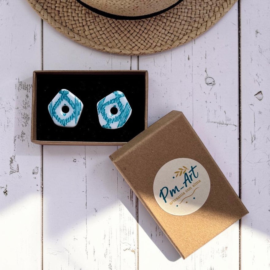 Pendientes artesanales turquesa con estampado tradicional mallorquín pintado a mano, presentados junto a su caja de embalaje bajo un sombrero de paja veraniego.